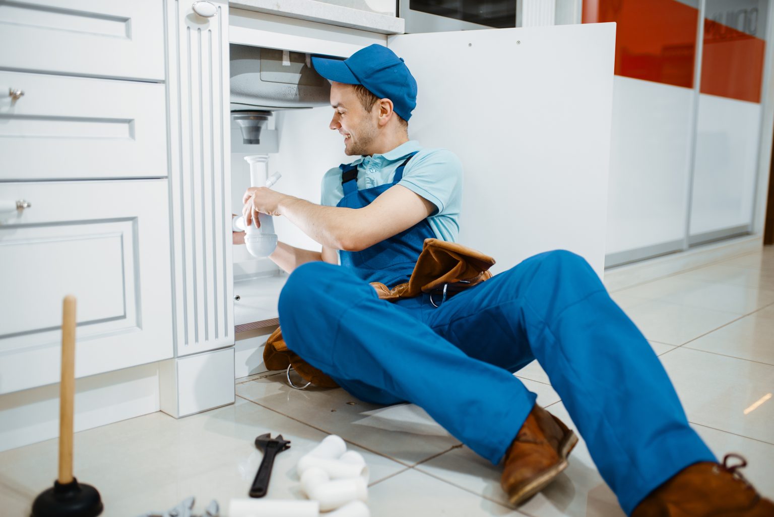 Smiling male plumber in uniform holds drain pipe in the kitchen. Handywoman with toolbag repair sink, sanitary equipment service at home