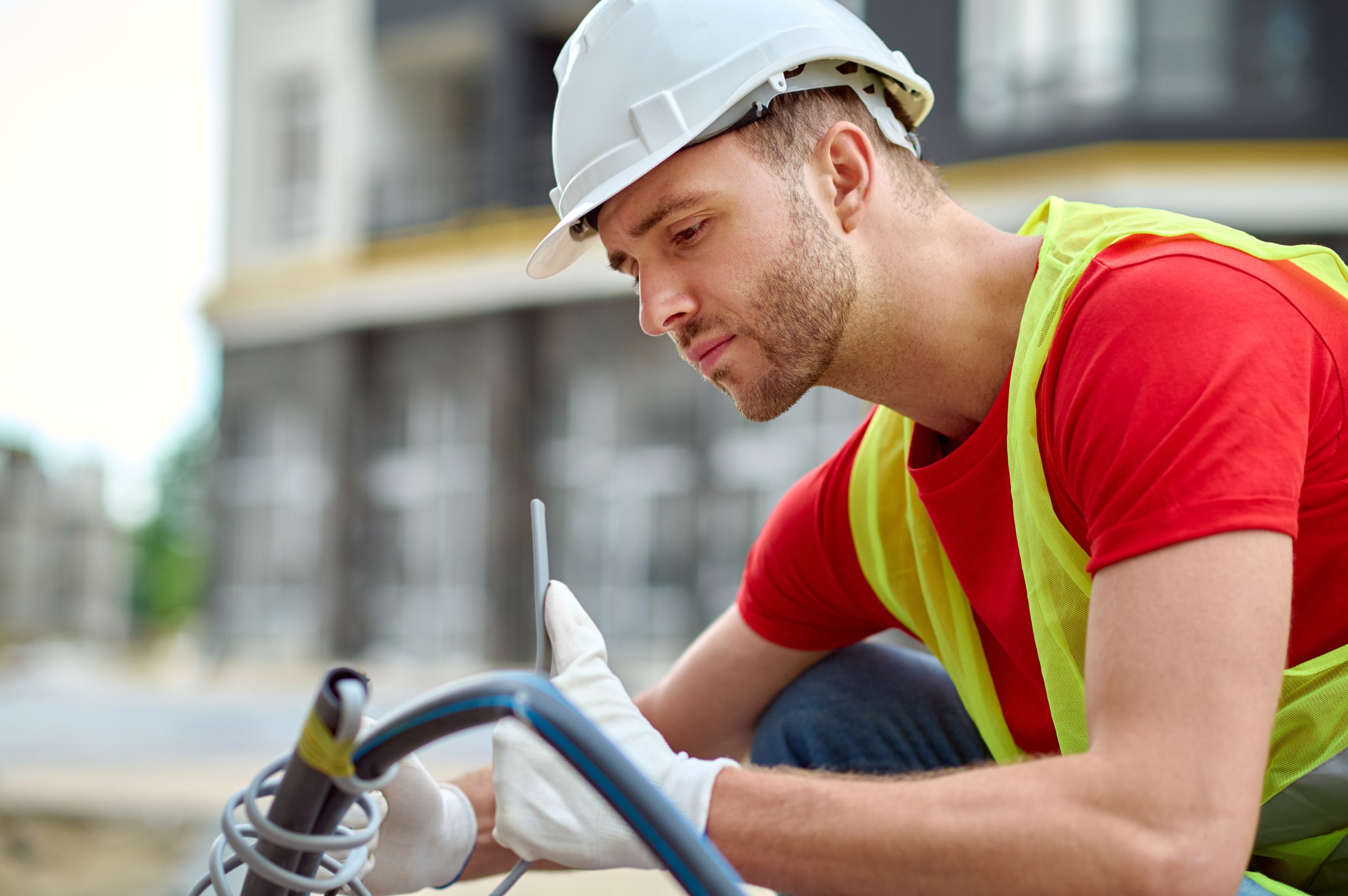 Adjustment. Young adult man in protective helmet gloves and bright vest crouching looking at tool sideways to camera at construction site